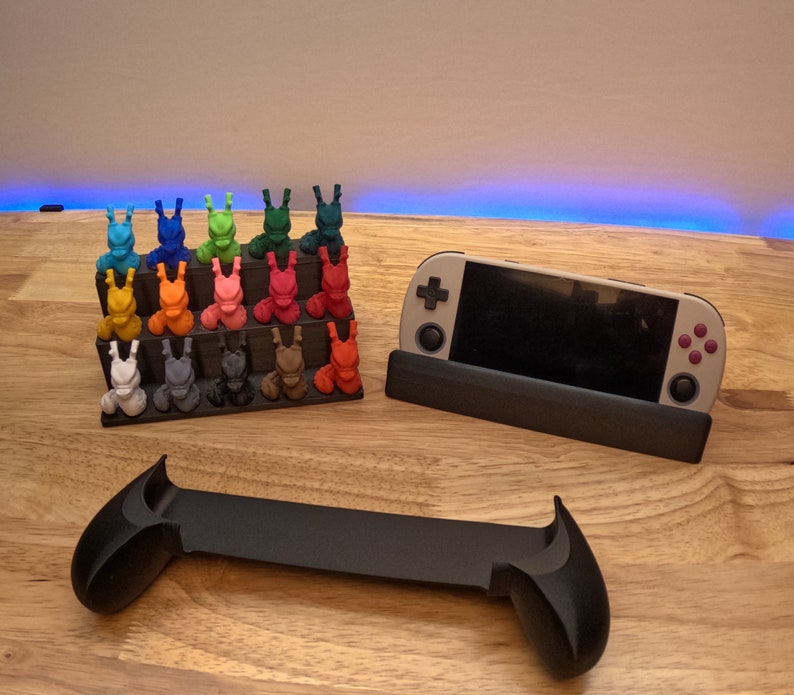 K&ouml;nnte beinhalten: Ein schwarzer St&auml;nder mit einer grauen Handheld-Spielkonsole, einem schwarzen Controller und einem mehrfarbigen 3D-gedruckten Displayst&auml;nder mit kleinen Figuren.