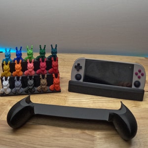 K&ouml;nnte beinhalten: Eine graue und schwarze Handheld-Spielkonsole mit einem St&auml;nder und einem Satz von 20 bunten 3D-gedruckten Figuren.