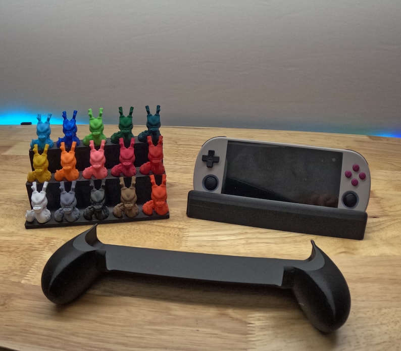 K&ouml;nnte beinhalten: Eine graue und schwarze Handheld-Spielkonsole mit einem St&auml;nder und einem Satz von 20 bunten 3D-gedruckten Figuren.