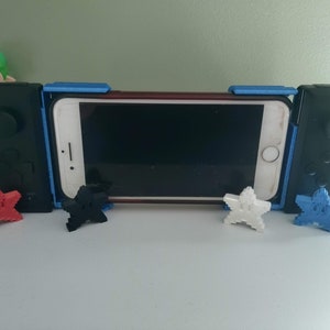 May include: A blue and black phone controller with a smartphone in the middle. The controller has two black buttons on each side and a blue plastic frame. There are five small star-shaped objects in different colours around the controller.