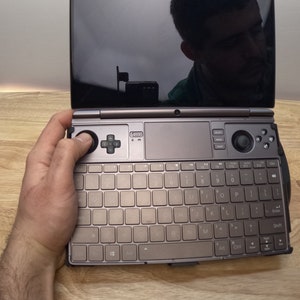 May include: A small, portable computer with a keyboard, trackpad, and a small screen. The computer is open and a person's hand is resting on the keyboard.