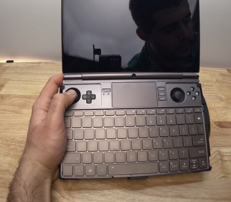 May include: A small, portable computer with a keyboard, trackpad, and a small screen. The computer is open and a person's hand is resting on the keyboard.