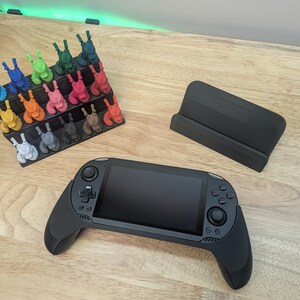 Puede incluir: Una consola de juegos port&aacute;til negra con una pantalla y dos mandos. La consola est&aacute; sobre una superficie de madera. Hay un soporte negro y una colecci&oacute;n de figuritas de colores en el fondo.