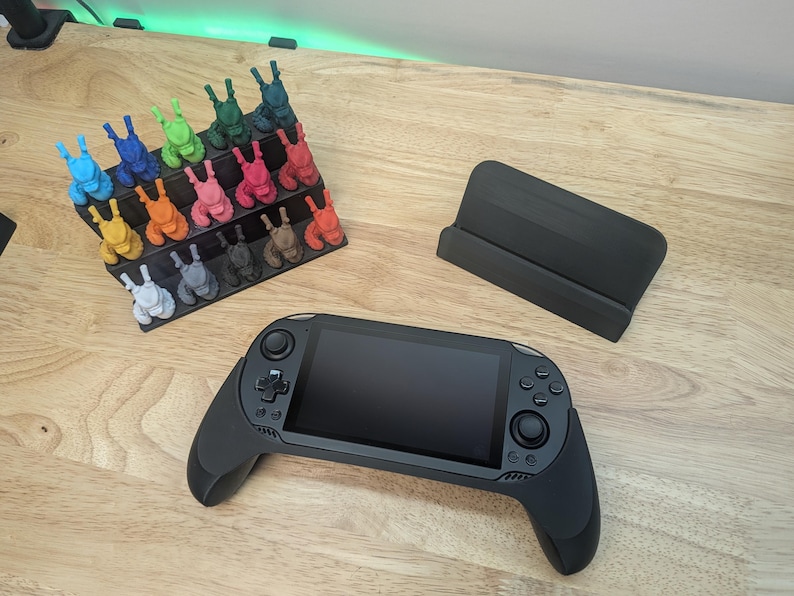 Puede incluir: Una consola de juegos port&aacute;til negra con una pantalla y dos mandos. La consola est&aacute; sobre una superficie de madera. Hay un soporte negro y una colecci&oacute;n de figuritas de colores en el fondo.