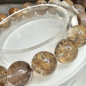 May include: A brown and clear quartz crystal bracelet with a natural, cloudy appearance. The bracelet is made of round beads and is displayed on a white stand.