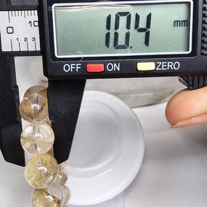 May include: A close-up of a digital caliper measuring the circumference of a bracelet made of clear quartz beads with brown inclusions. The caliper reads 10.4 mm.