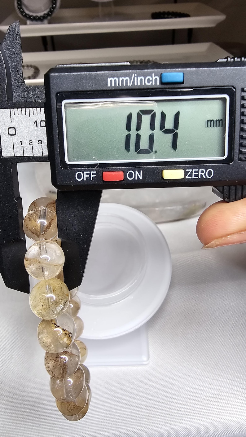May include: A close-up of a digital caliper measuring the circumference of a bracelet made of clear quartz beads with brown inclusions. The caliper reads 10.4 mm.