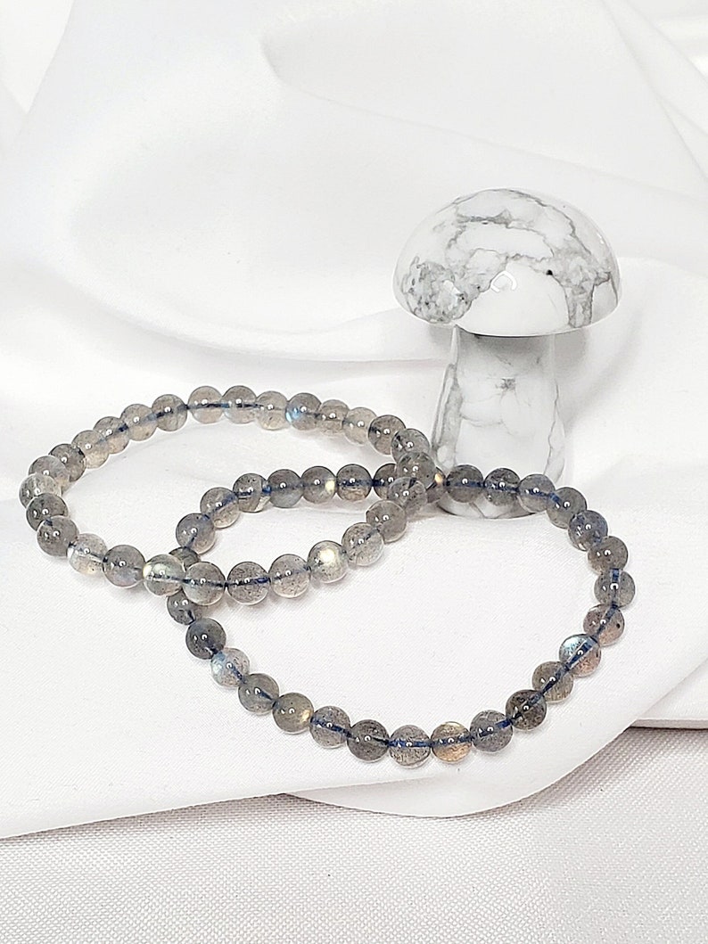 May include: Two gray labradorite beaded bracelets on a white surface. The bracelets are made of round beads and are stacked on top of each other.