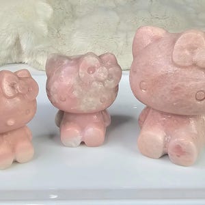 Puede incluir: Tres figuras de piedra rosa de un gato con un lazo, en diferentes tamaños. Las figuras de gato tienen un aspecto liso y pulido y están de pie. El color es rosa suave con vetas blancas. Se exhiben sobre una superficie blanca.