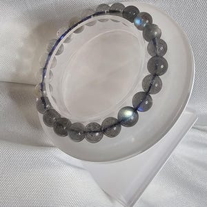 May include: A gray and blue labradorite gemstone bracelet on a white display stand. The bracelet is made of round beads and has a iridescent sheen.