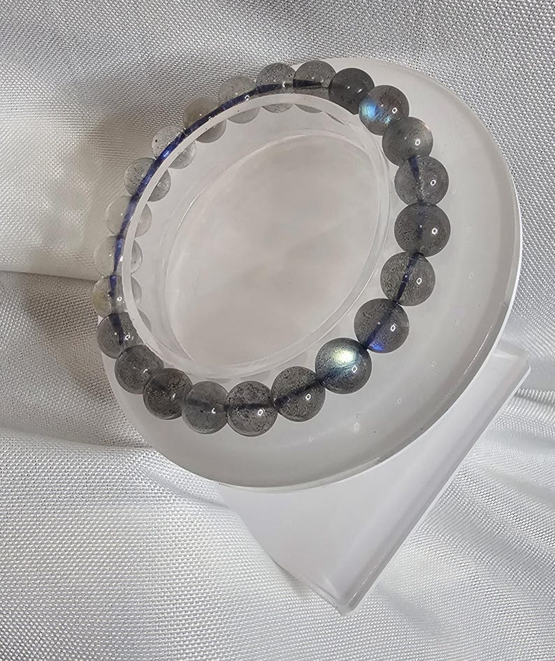 May include: A gray and blue labradorite gemstone bracelet on a white display stand. The bracelet is made of round beads and has a iridescent sheen.