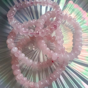 May include: Three pink rose quartz beaded bracelets stacked on a iridescent background. The bracelets are made of smooth, round beads.