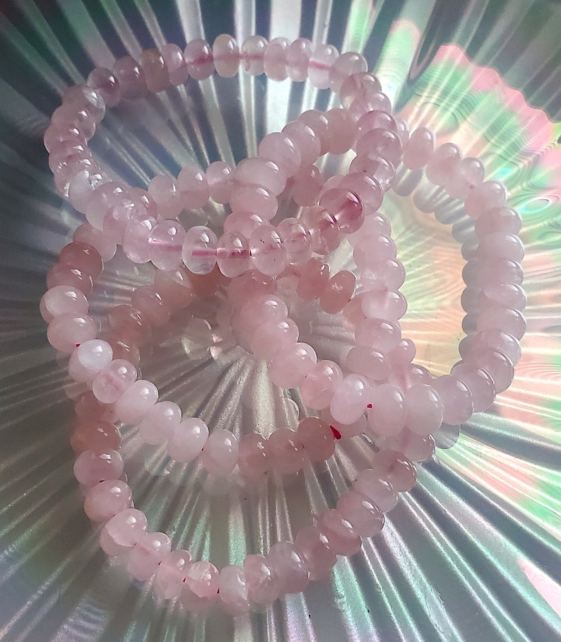 May include: Three pink rose quartz beaded bracelets stacked on a iridescent background. The bracelets are made of smooth, round beads.
