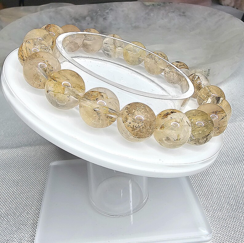 May include: A bracelet made of golden brown, translucent, round beads with a clear, crystalline texture. The beads are arranged in a circle on a white display stand.