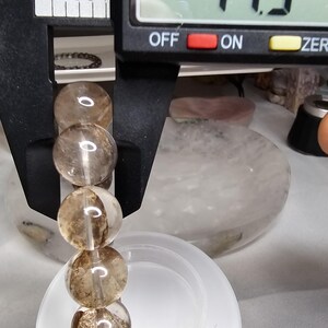 May include: A close-up of a brown and clear crystal bead bracelet being measured with a digital caliper. The caliper reads 1.15 inches.