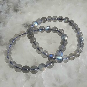 May include: Two gray labradorite gemstone bracelets with iridescent blue and green hues. The bracelets are made of round beads and are stacked on top of each other.