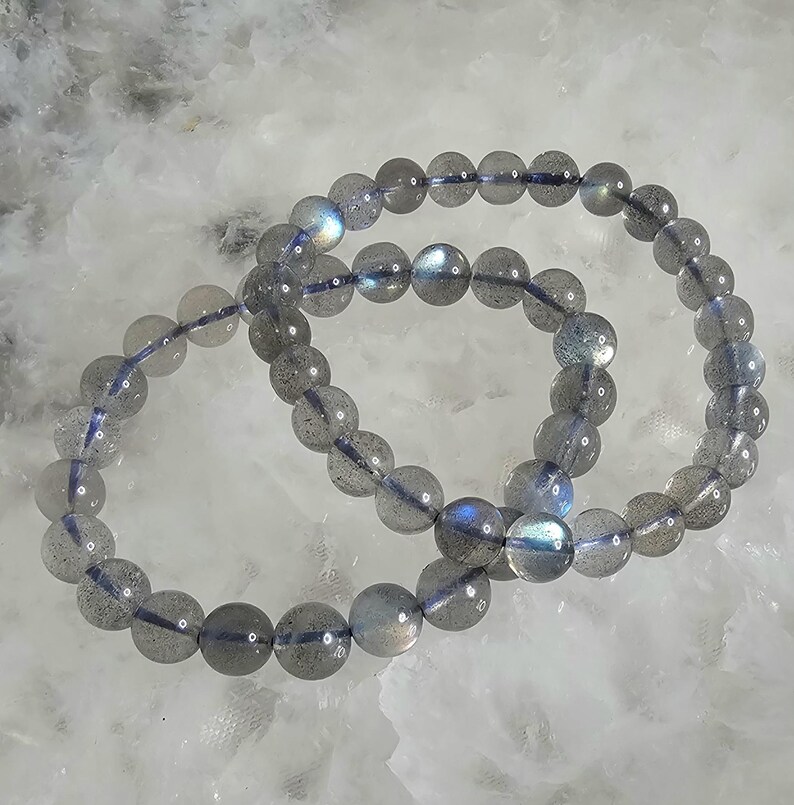 May include: Two gray labradorite gemstone bracelets with iridescent blue and green hues. The bracelets are made of round beads and are stacked on top of each other.