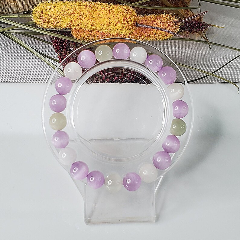 May include: A bracelet made of purple, white, and green gemstone beads. The bracelet is on a clear plastic stand.
