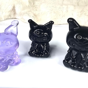 Puede incluir: Tres figuras talladas con forma de personaje de dibujos animados. Una es de color púrpura translúcido y las otras dos son de color negro brillante. Las figuras tienen orejas grandes, caras redondas y están sentadas. Se exhiben sobre una superficie blanca.