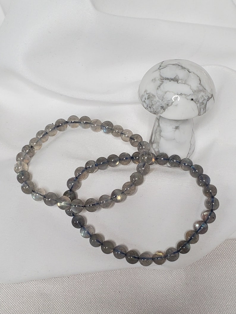 May include: Two gray labradorite gemstone bracelets with a white mushroom figurine in the background.