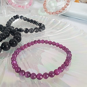 May include: A purple gemstone bracelet with round beads. The bracelet is on a white, iridescent surface.