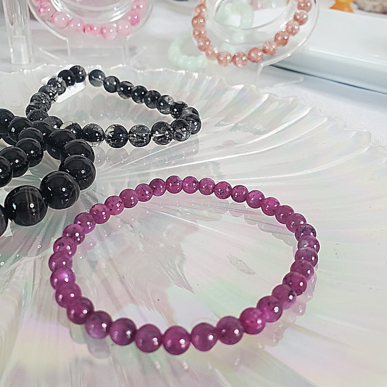 May include: A purple gemstone bracelet with round beads. The bracelet is on a white, iridescent surface.