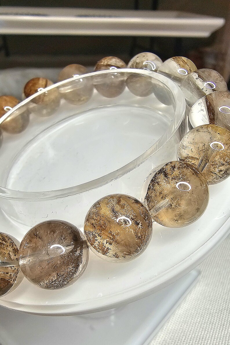 May include: A brown and clear crystal bracelet with a natural, cloudy appearance. The bracelet is made of round beads and is displayed on a white stand.