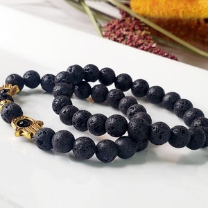 May include: Two black lava stone bracelets with gold hamsa hand charms. The bracelets are stacked on top of each other.