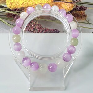 May include: A bracelet made of purple, white, and green beads. The beads are round and have a smooth, polished finish. The bracelet is displayed on a clear plastic stand.
