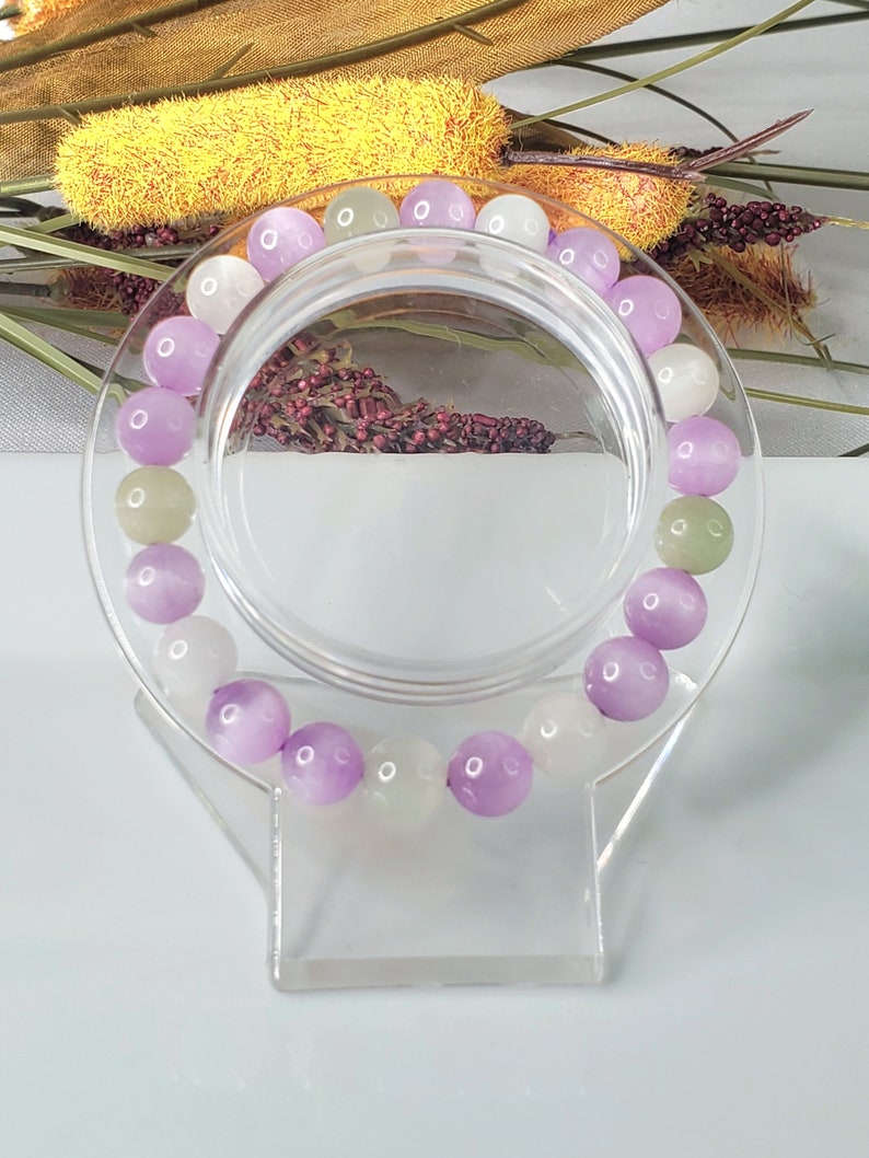 May include: A bracelet made of purple, white, and green beads. The beads are round and have a smooth, polished finish. The bracelet is displayed on a clear plastic stand.