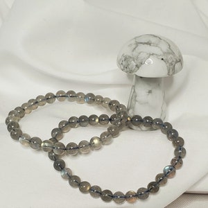 May include: Two gray labradorite gemstone bracelets with iridescent blue and gold flecks. The bracelets are stacked on top of each other and are lying on a white surface.