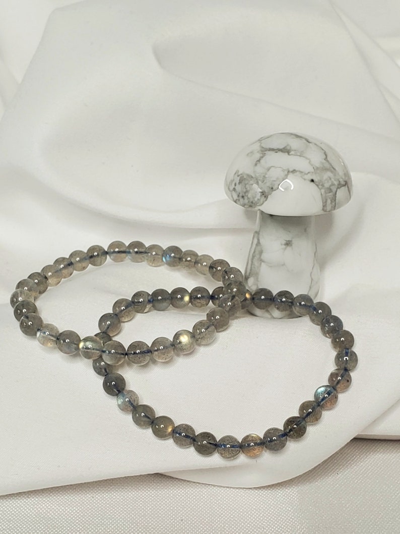 May include: Two gray labradorite gemstone bracelets with iridescent blue and gold flecks. The bracelets are stacked on top of each other and are lying on a white surface.