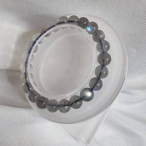 May include: A gray labradorite gemstone bracelet with iridescent blue and green hues. The bracelet is made of round beads and is displayed on a white stand.