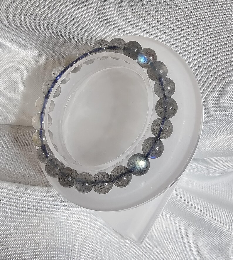 May include: A gray labradorite gemstone bracelet with iridescent blue and green hues. The bracelet is made of round beads and is displayed on a white stand.