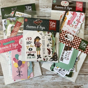 May include: Several packages of die-cut ephemera cardstock pieces, including frames and tags, are displayed. The packages feature various themes, such as pirates, farm life, and adventure. The color palette includes green, red, and pink.