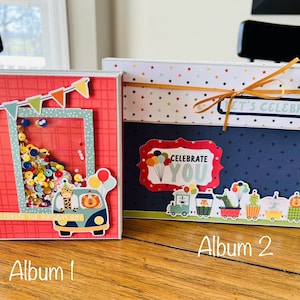 May include: Two handmade photo albums with a birthday theme. The first album is red with a shaker window and a cartoon van with animals. The second album is blue with a star pattern and a banner that says "Let's Celebrate!"