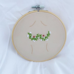 May include: Embroidered hoop art with a floral design in shades of green, white, and pink. The design is centered in the hoop and is surrounded by a beige fabric.