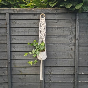 May include: A white macrame plant hanger with a wooden ring at the top, hanging from a black wooden fence. A green plant is in a white pot inside the hanger.