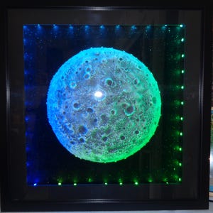 Könnte beinhalten: Eine 3D-Mondskulptur mit einer strukturierten Oberfläche, beleuchtet von blauen und grünen LED-Lichtern in einem schwarzen Rahmen.