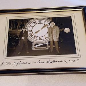 May include: A framed black and white photograph featuring two figures standing in front of a large clock face. The text "To Marty Partners in time September 5, 1885" is written in black ink on the white matting below the photograph.