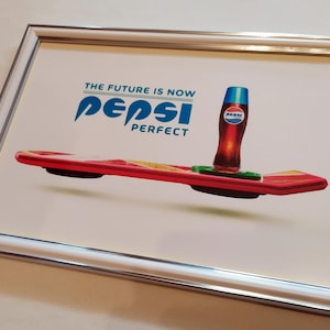 Puede incluir: Impresión enmarcada con una botella de Pepsi Perfect sobre una tabla flotante roja y amarilla. La imagen incluye el texto "THE FUTURE IS NOW" y "Pepsi PERFECT" en azul y blanco. El marco es plateado.