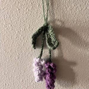May include: A crocheted lavender flower with green leaves hanging from a string. The flower is made of purple and white yarn.