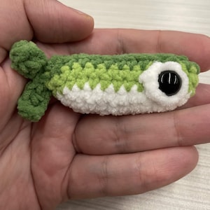 May include: A small, handmade crochet fish toy featuring a gradient of green yarn on the top and white yarn on the bottom. The fish has a large, round, black plastic safety eye surrounded by a white crocheted border and a textured green tail fin.