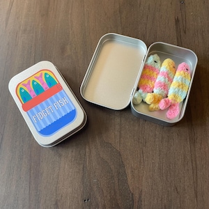 May include: A rectangular metal tin with a lid featuring a graphic of colorful fish and the text FIDGET FISH. Beside it, an open metal tin contains three small, soft, plush fish toys in pastel yellow, pink, blue, and white striped patterns.