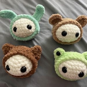 May include: Four handmade crochet plushies in the shape of animals. There is a bunny, two bears, and a frog. Each plushie has a white face with black eyes and is wearing a colored hood. The bunny is mint green, the bears are brown, and the frog is light green.