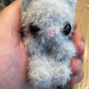 May include: A small, fuzzy, light gray plush animal figurine with a soft, textured appearance. It features two large, round, glossy black eyes and a tiny, pale pink stitched nose. The creature has pointed ears and a compact, seated posture.