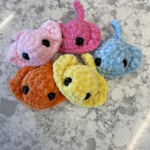 May include: Five small, plush, crocheted stingray toys in light pink, hot pink, light blue, orange, and yellow. Each soft, textured creature features two round, black plastic safety eyes and a small, pointed tail extending from the top center.