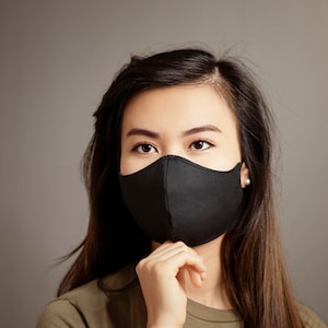 Op de afbeelding: Een vrouw met een zwart mondkapje en een groen T-shirt. Ze kijkt naar de camera.