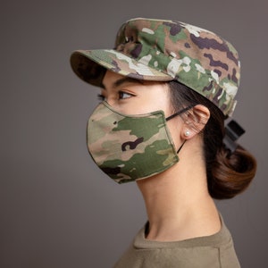 Op de afbeelding: Een vrouw draagt ​​een groen camouflagegezichtsmasker en een groene camouflagemilitaire pet. Ze draagt ​​een groen T-shirt.