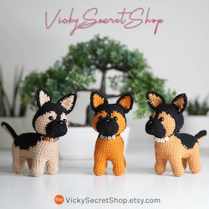 Op de afbeelding: Drie gehaakte Duitse herderhonden beeldjes in bruin, zwart en oranje. Elke hond heeft een halsband met kralen. De honden worden tentoongesteld voor een groene plant en twee cactussen.